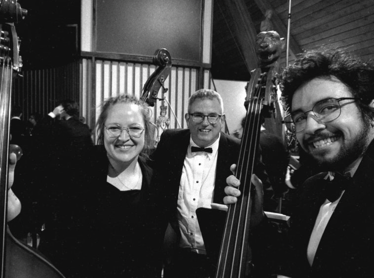 Three musicians, one being gabrielle matin, smiling warmly during a performance, holding double basses. The black-and-white image captures a moment of camaraderie and joy in a concert setting, with the intricate wooden details of the instruments adding a touch of elegance.